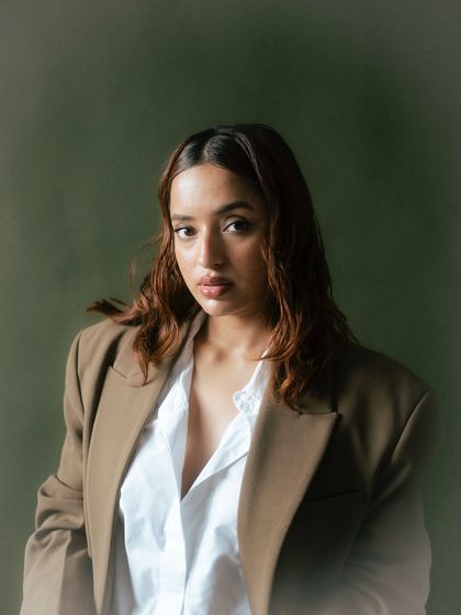 A direct and engaging portrait against a solid olive background. The simple composition and the model's strong eye contact make this a powerful and classic fashion image.