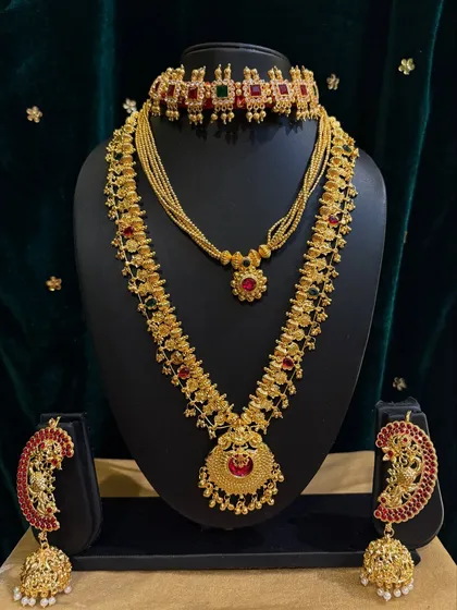 This rental set features a stunning choker and a long haar with peacock-shaped earrings, all studded with red and green stones.