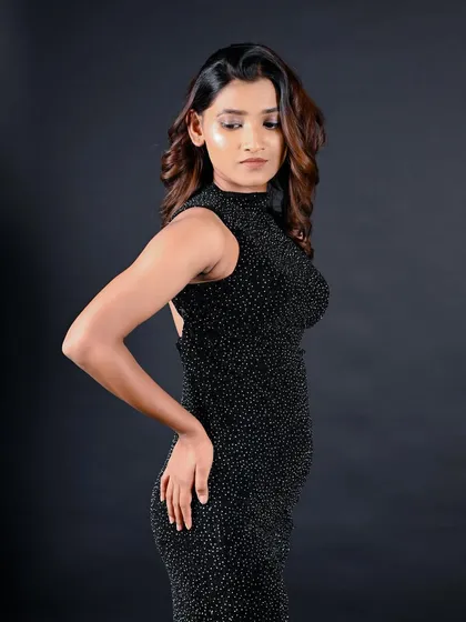 A three-quarter portrait of a model in a sparkling black dress against a dark backdrop. The lighting is sculpted to highlight her form and the dress texture.