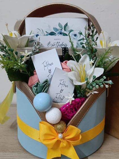 A beautiful birthday basket with a blue and yellow theme. It's adorned with white lilies and contains scrunchies and other small gifts, perfect for a sister or friend.