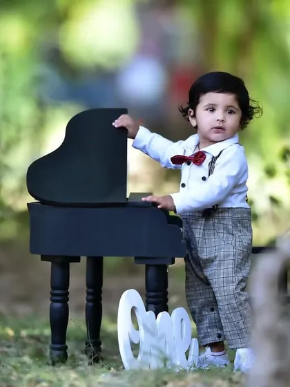 A full shot of the little pianist theme, with the one year old boy standing proudly next to his mini piano in a beautiful park setting.