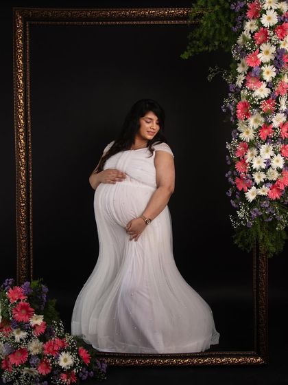 A stunning portrait using a large frame decorated with fresh flowers. The mom-to-be in a classic white gown looks absolutely angelic in this luxurious setup.