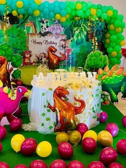 A prehistoric adventure with this fun dinosaur-themed birthday party. The setup includes a lush green backdrop, various dinosaur cutouts and foil balloons, and a balloon-wrapped cake table.