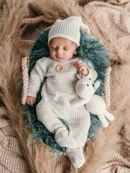 A sleeping newborn in a cozy, textured white romper and matching hat, holding a small knitted toy. This boho-inspired shot uses natural textures like pampas grass for a warm, earthy feel.