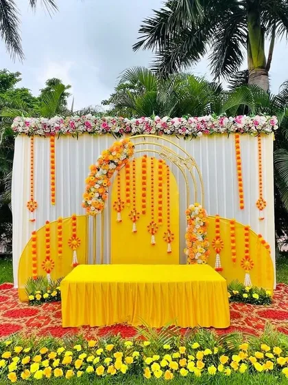 A modern and stylish Haldi stage setup for an outdoor event. This design uses a combination of white drapes, geometric golden arches, and marigold flower arrangements for a fresh and contemporary look.