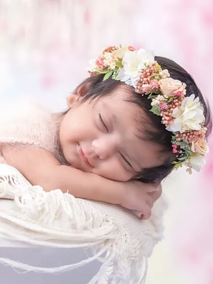 A gentle, sleepy smile is one of the most precious moments to capture. This close-up highlights the baby's peaceful expression, framed by a delicate floral headband against a soft pink background.