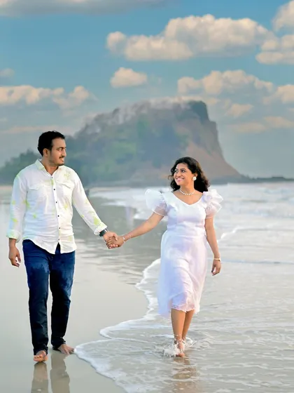 A candid moment of a couple walking hand-in-hand along the water's edge. The bright, sunny day and the distant coastal mountain create a beautiful, natural setting for this pre-wedding photograph.
