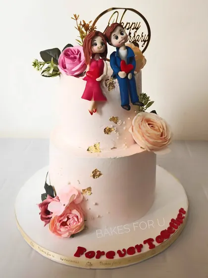 "Forever Together." A romantic two-tier anniversary cake with custom fondant figures of the couple, surrounded by fresh flowers and delicate gold leaf.