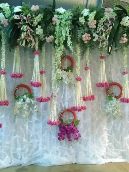 A beautiful floral backdrop made of white and pink hanging flower strings (kaleeras) and orchid arrangements against a white ruffled curtain, perfect for a home engagement.