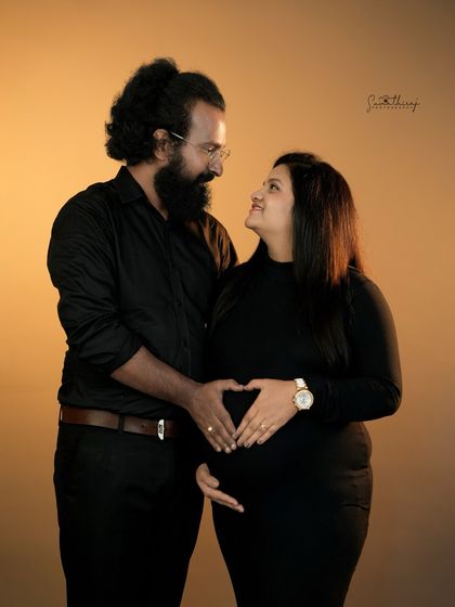 A loving couple portrait against a warm, golden-lit background. Their hands form a heart on the baby bump, symbolizing the love that surrounds their growing family.