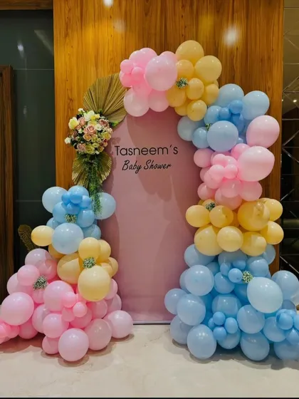 An elegant and modern baby shower backdrop. This setup uses a beautiful pastel color palette of pink, blue, and yellow balloons, accented with dried palm leaves and fresh flowers for a sophisticated look.
