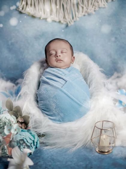 A baby boy swaddled in blue, resting in a soft white nest surrounded by blue flowers and candles for a serene, dreamy look.
