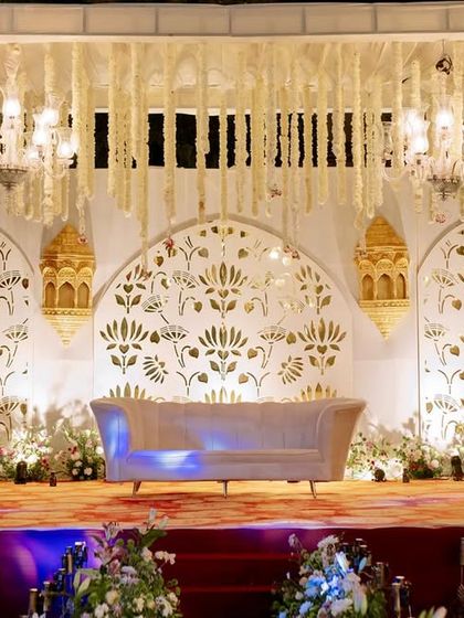 An elegant white and gold stage for a traditional ceremony, featuring intricate jaali work, hanging floral strings, and ornate lanterns.