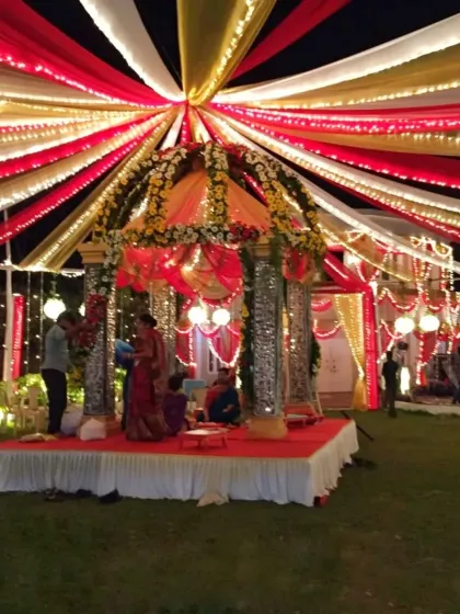 A vibrant wedding mandap under a canopy of red, white, and yellow drapes, with decorated pillars and ample lighting for a nighttime ceremony.