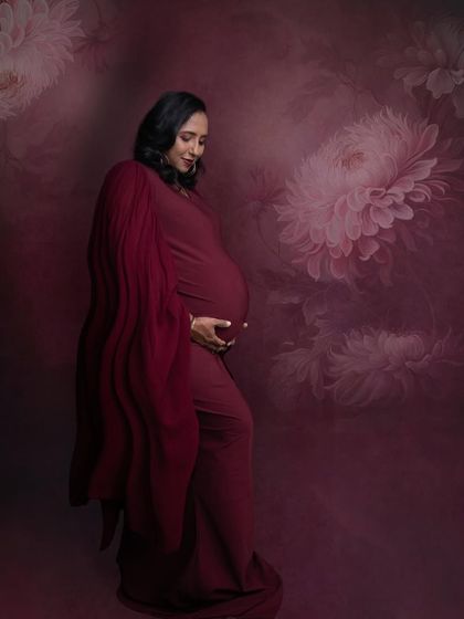A soft and gentle portrait. The mom-to-be cradles her bump in a beautiful red gown, set against a romantic, painted floral background.