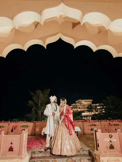 A wide angle view of the couple standing under a traditional archway, with the majestic palace in the distance. This shot establishes the scale and beauty of a destination palace wedding.