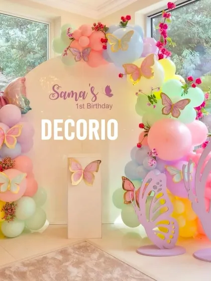 A full view of the pastel butterfly-themed first birthday party. The setup includes an arched backdrop, butterfly cutouts, and a soft, multi-colored balloon garland.