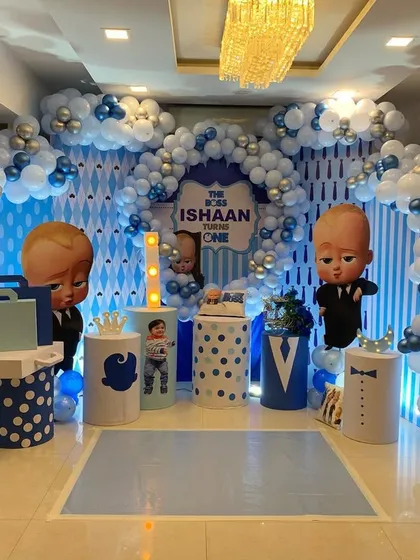 A 'Boss Baby' themed first birthday party. The blue, white, and silver color scheme is used across the balloon arch, backdrop, and themed props like briefcases and letter blocks spelling 'ONE'.