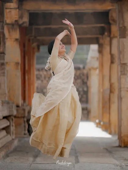 A bride-to-be, dressed in an elegant cream saree, leaps with joy in a stone temple hallway. This captures her personality and the happiness of the moment.