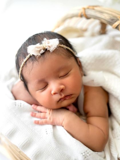 A close-up portrait of a baby sleeping peacefully in a basket, wrapped in a soft white blanket. The focus is all on her sweet, calm face.