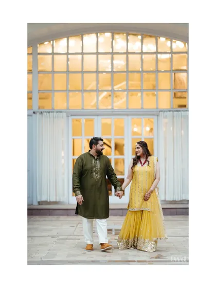A sweet, candid moment during a mehndi celebration. The couple holds hands, sharing a look that speaks of shared joy and anticipation for the journey ahead.