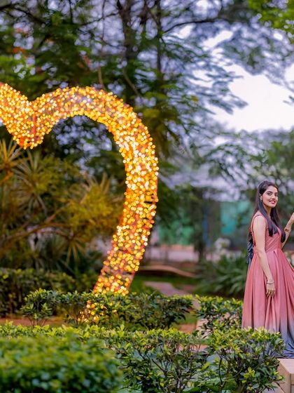 A couple poses by the illuminated heart prop and stone gazebo at dusk, a magical garden setting.