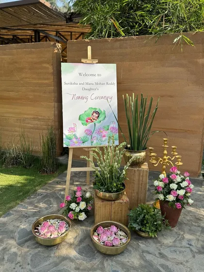 A beautiful entrance setup with a hand-painted sign, brass urlis filled with lotus petals, and rustic wooden pedestals.