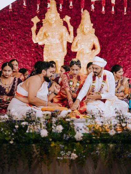 A wide shot of the wedding ceremony, showing the couple surrounded by flowers, deities, and family.