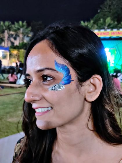 A smiling client with a blue glitter butterfly, enjoying an outdoor festival.