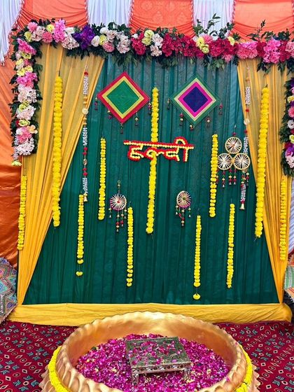 A traditional Haldi setup inside a tent. The backdrop of green and yellow drapes is decorated with flowers and hangings, and the large urli is filled with rose petals for the ceremony.