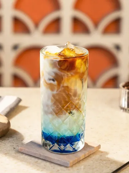 A layered iced coffee with blue syrup at the bottom, transitioning to milky white and rich brown espresso. The warm sunlight and patterned background give this shot a cozy cafe vibe.