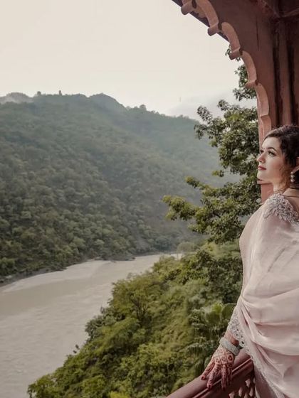 The bride-to-be stands on a balcony overlooking a river winding through lush green mountains. The flowing saree adds a sense of movement and grace to this breathtaking scenic portrait.