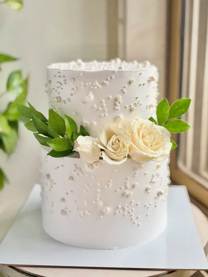 A vision in white, this two-tiered cake is the definition of timeless elegance. The clean buttercream finish is adorned with scattered edible pearls and fresh white roses, making it a perfect centerpiece for a sophisticated roka or wedding celebration.