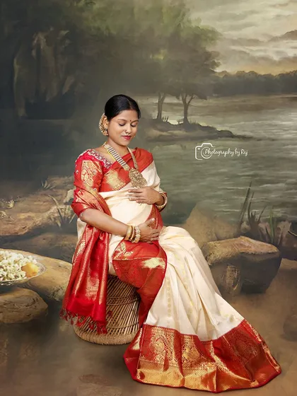 A peaceful studio portrait of a mother-to-be in a traditional red and white saree, seated against a painted backdrop.