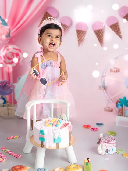 A candid moment of delight during a first birthday cake smash. Dressed in a pastel gown amidst a sweet-themed backdrop, this little one enjoys her special treat, creating a perfect memory.