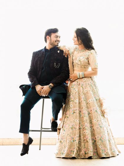 A bright and cheerful studio portrait against a clean white background. The playful interaction between the seated groom and standing bride creates a fun and relaxed pre-wedding photo.