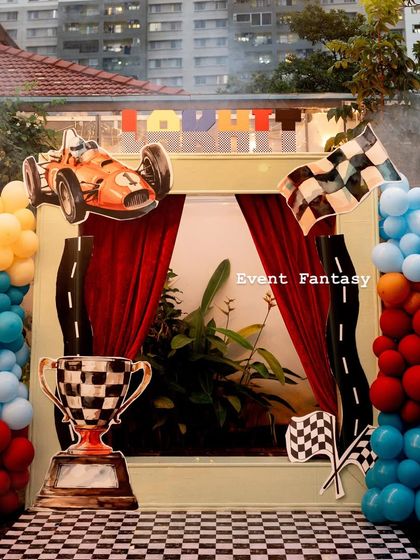 A dynamic photo booth for Lakhit's "Fast One" racing party. The setup includes a vintage race car cutout, a large trophy, and a checkered floor, all framed by red curtains.