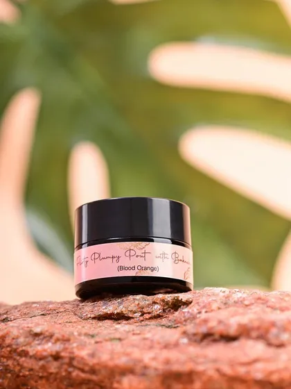 A close-up of a Pure by Priyanka lip tint on a textured rock, with a large leaf in the background. The composition is simple yet effective at creating a natural, tropical vibe.
