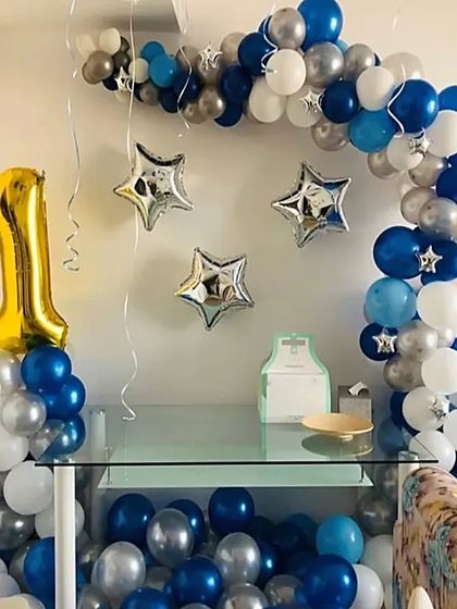 A simple yet charming first birthday setup at home. It features a blue, white, and silver balloon arch over a glass table and a large number '1' foil balloon.