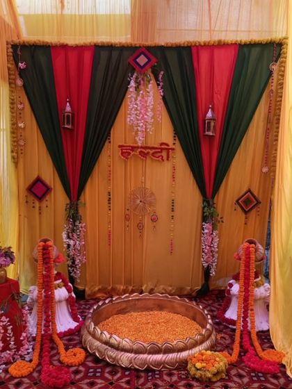 A traditional Haldi setup with a large brass urli filled with marigold petals. The backdrop is created with red, yellow, and green drapes, perfect for a vibrant home ceremony.