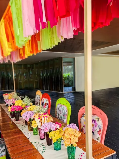 A long table setup for the Haldi event, with a colorful tassel canopy overhead and a line of vibrant floral arrangements in quirky vases.