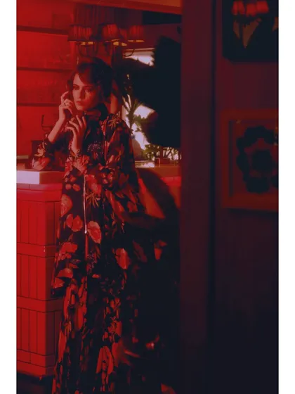A vertical composition from 'The Wait', emphasizing the length of the floral coat and the atmospheric, shadow-filled room. Every element is styled to contribute to the narrative of waiting.