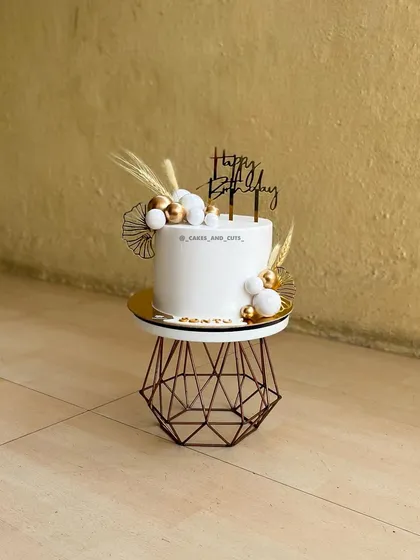 A modern and aesthetic white cake decorated with dried wheat, pampas grass, and gold and white spheres.