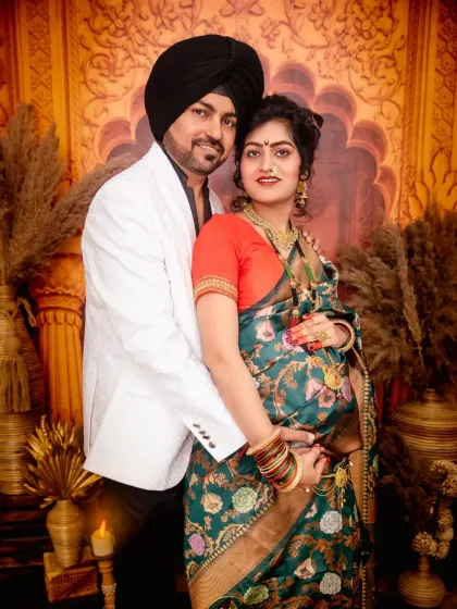 A joyful couple's portrait celebrating their pregnancy in traditional attire. The partner stands behind his wife, who is dressed in a beautiful green saree, both smiling at the camera.