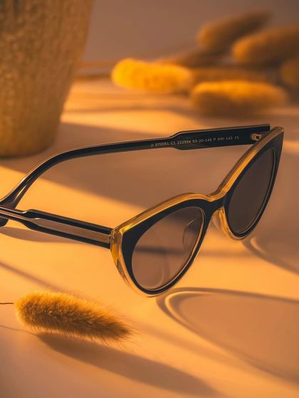 A still-life product shot of John Jacobs sunglasses, styled with dried flowers on a warm, sunny surface to evoke a winter-sun aesthetic.