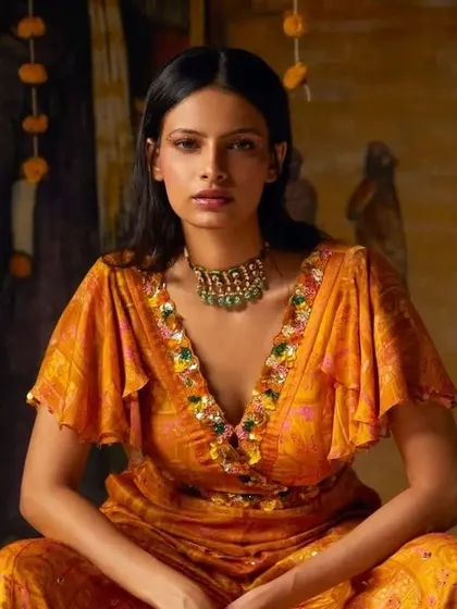 A seated portrait from a designer campaign, showcasing a rich, warm-toned makeup look. The makeup complements the orange and gold hues of the outfit, creating a cohesive and luxurious feel for the brand's lookbook.