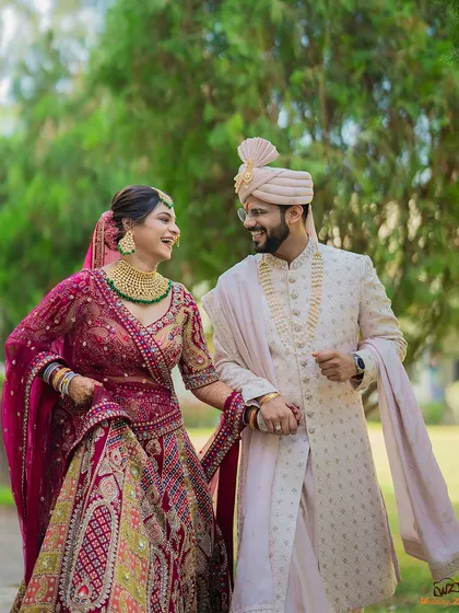 A candid, joyful moment of a couple walking hand-in-hand during their Gujarati wedding celebrations, showcasing their vibrant red and cream outfits.