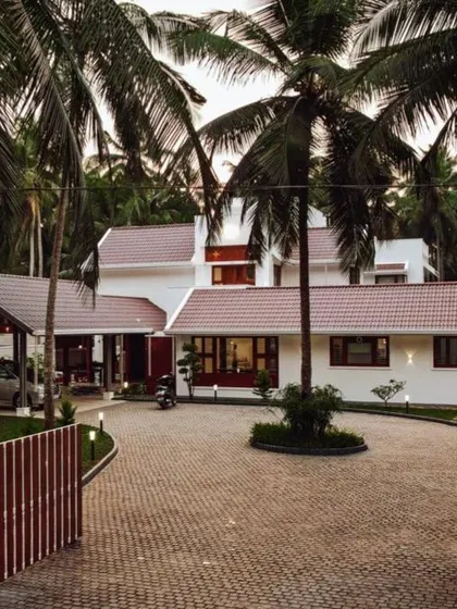 The entrance to the 'Coconut Cove' residence provides a ground-level perspective of how the contemporary Kerala home design integrates with the landscape. The paved driveway and strategic lighting guide you into a tranquil living space.