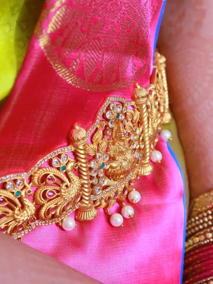 A close-up of a traditional gold Vaddanam (waist belt) with a goddess motif, worn over a bright pink silk saree.