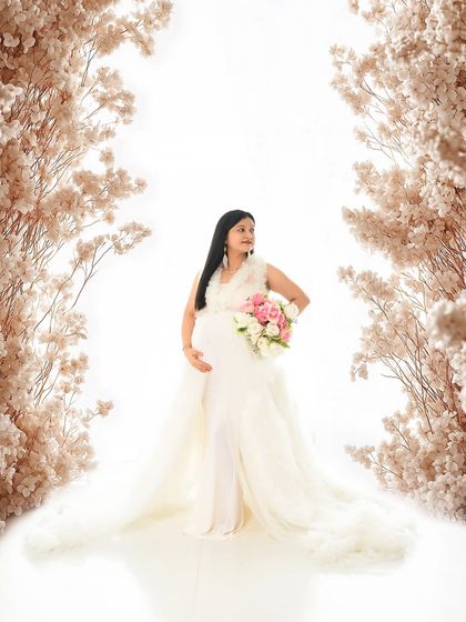 A breathtaking solo portrait of the mom-to-be in a white gown, standing between two large white floral arrangements. The high-key lighting creates an ethereal, heavenly glow.